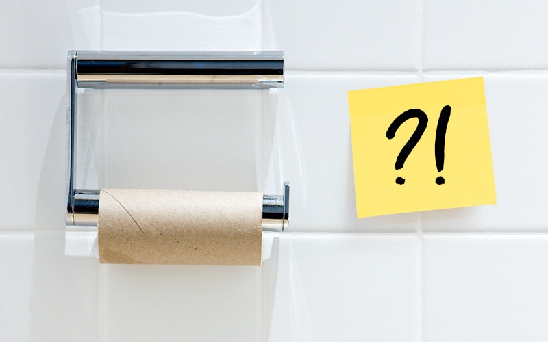 Leerer Halter für Kleinrollen-Toilettenpapier mit einem Post-it daneben, auf dem eine Frage und ein Ausrufezeichen stehen
