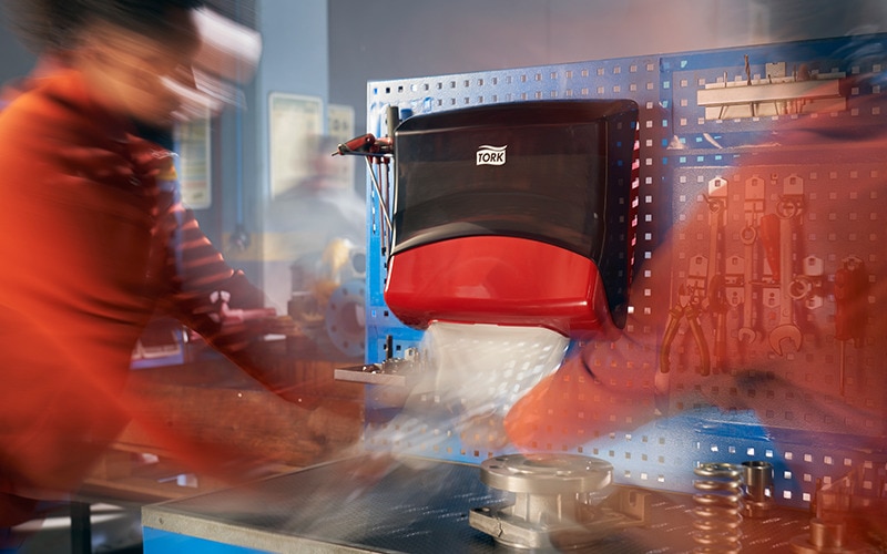 Una imagen a cámara rápida muestra a dos operarios de fábrica extrayendo toallas de papel de un dispensador Tork. 