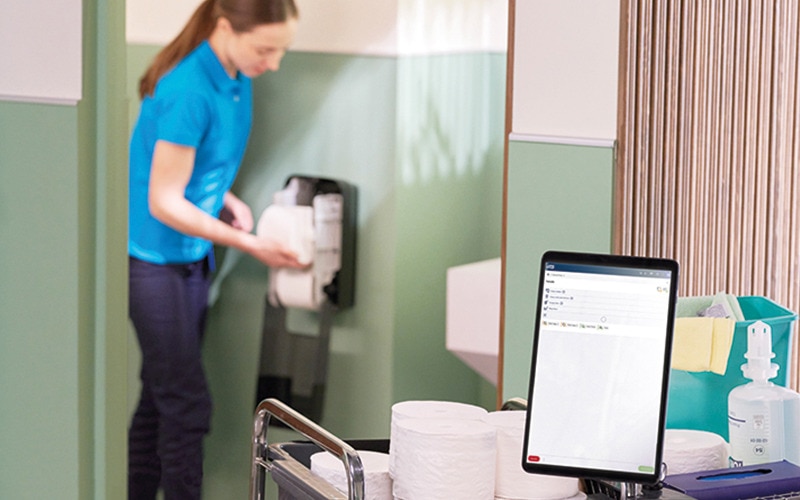 Close-up van een tablet op een schoonmaaktrolley en een schoonmaker die een toiletpapierdispenser vult