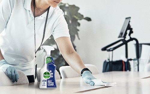 A cleaner wiping a table, on the table there is a bottle of Tork Dettol Antibacterial Multipurpose Cleaner