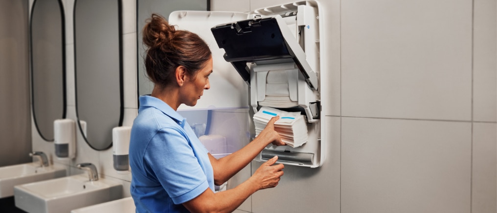 Person fyller på en väggmonterad pappersdispenser, PeakServe Automatic, i en offentlig toalettmiljö