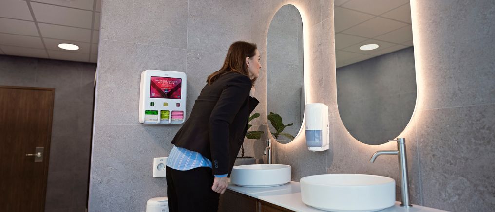 Ei person lener seg fram mot eit vaskeservant og spegel på eit baderom, med ein mensbeskyttingsdispenser montert på veggen ved sida av