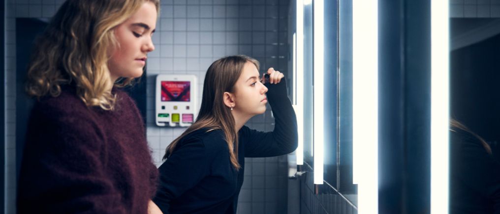 Två elever står vid en spegel i ett badrum, en justerar sitt ansikte medan den andra står bredvid. En mensskyddsdispenser syns på väggen i bakgrunden.