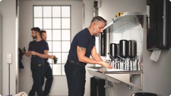 Person washing hands at a sink in a restroom equipped with multiple dispensers, with others standing in the background Person washing hands at a sink in a restroom equipped with multiple dispensers, with others standing in the background
