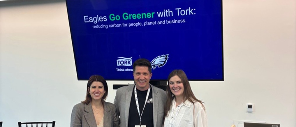 Three people stand in front of a large presentation screen displaying the text “Eagles Go Greener with Tork: reducing carbon for people, planet and business” alongside the Tork logo and the Philadelphia Eagles logo