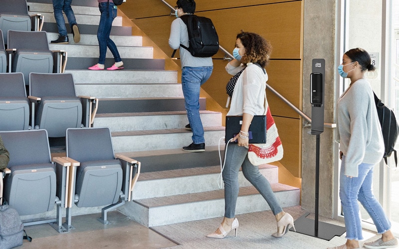 Étudiants portant des masques de protection entrant dans la salle de conférence comportant un pied pour distributeur de mousse soin de la peau Tork à l’entrée 