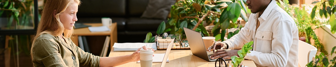 A woman and a man are sitting at a table working on their laptops with a cup of coffee each, in the background a black couch and some green plants