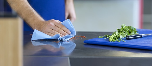 A hand is holding a blue cloth and is wiping some spills, in front a blue cutting board with fresh coriander and a knife