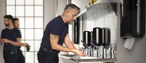 a man is washing his hands in a restroom with a long sink, several soap dispensers and paper towel dispensers, in the background to the left two more men