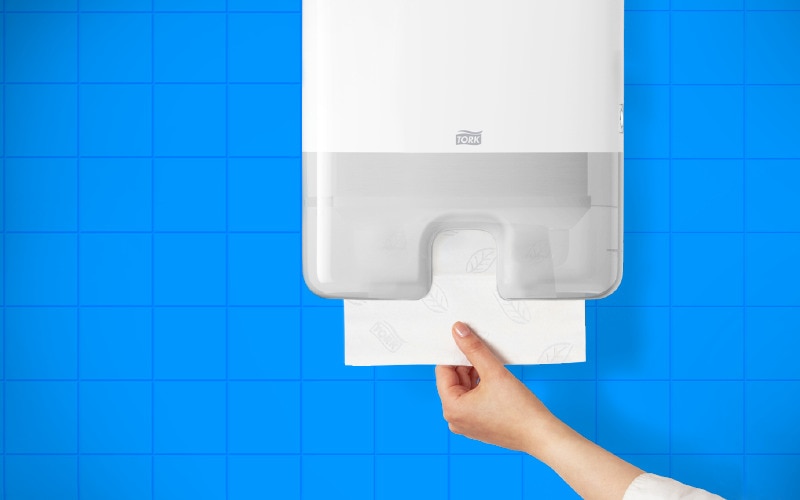 A hand pulling paper hand towel from a white dispenser, background is bright blue