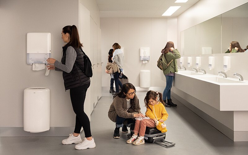 Kvinnor i ett offentligt toalettutrymme med Tork dispensrar för handdukar och tvål.