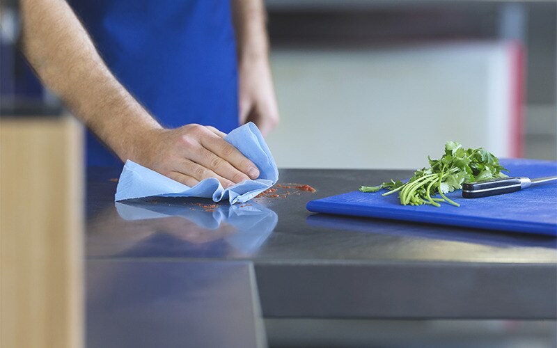 a hand is holding a blue cloth and is wiping some spills; in front, a blue chopping board with fresh coriander and a knife