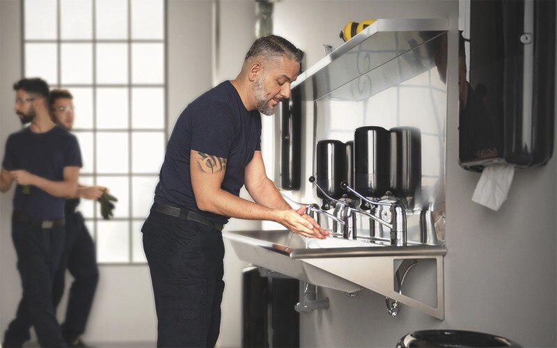 a man is washing his hands in a restroom with a long sink, several soap dispensers and paper towel dispensers, in the background to the left two more men