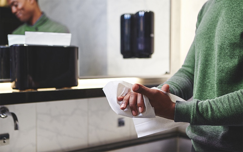 a man is wiping his hands with a paper towel; in the background, he’s in the mirror and there is also a paper towel dispenser and a soap dispenser