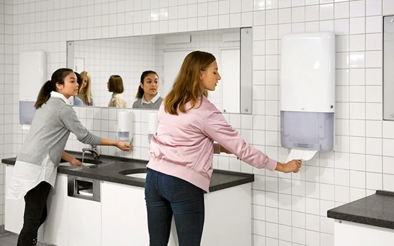 A woman washes her hands and another woman uses a paper towel dispenser