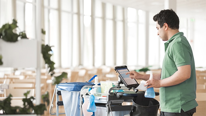 Een schoonmaker gebruikt een tablet naast een schoonmaakkar op een lichte, open locatie.