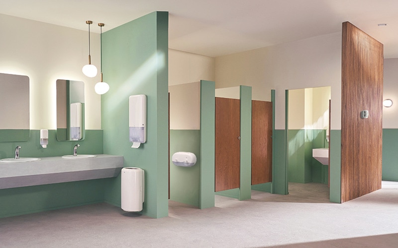 Modern public restroom with light green walls and wooden stall doors, featuring two sinks with mirrors, and soap dispensers. A paper towel and airfreshener dispensers are mounted on the wall.