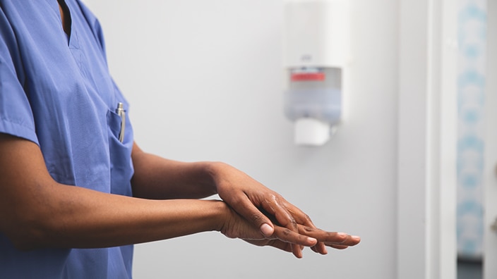 Person in a clinical environment applying hand sanitiser to hands, with a wall-mounted dispenser visible behind.