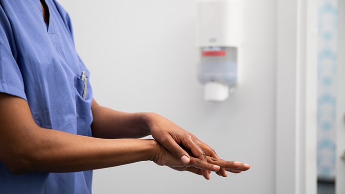 Person in a clinical environment applying hand sanitiser to hands, with a wall-mounted dispenser visible behind.
