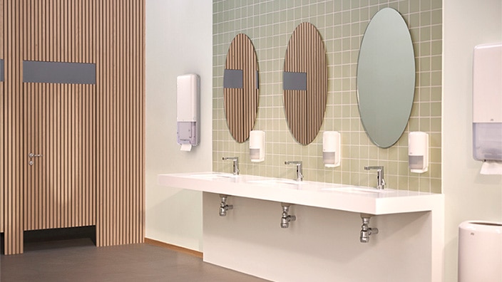 Et toilet med tre håndvaske, ovale spejle og en blanding af træ og grønne fliser, inklusive dispensere med papirhåndklæder og sæbe