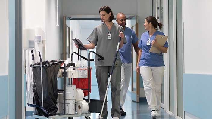 Hospital cleaner pushing a cleaning cart with ipad and staff in scrubs with clipboards. Hospital cleaner pushing a cleaning cart with ipad and staff in scrubs with clipboards.
