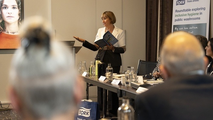 A speaker stands at the front of a meeting room addressing participants seated around a conference table A speaker stands at the front of a meeting room addressing participants seated around a conference table
