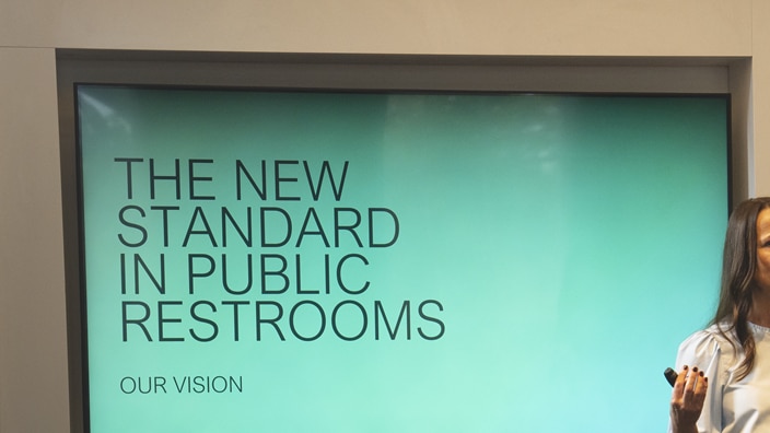 A woman is giving a presentation, a screen displaying the text ‘The new standard in public washrooms – our vision’. A woman is giving a presentation, a screen displaying the text ‘The new standard in public washrooms – our vision’.