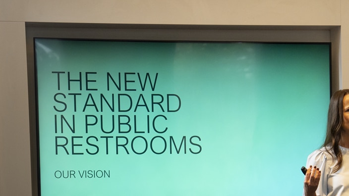 A woman is giving a presentation, a screen displaying the text ‘The new standard in public washrooms – our vision’. A woman is giving a presentation, a screen displaying the text ‘The new standard in public washrooms – our vision’.