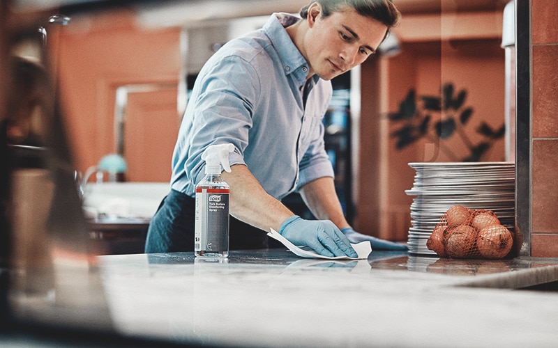 a man is cleaning a surface in a restaurant kitchen with Tork disinfecting spray and a wiper