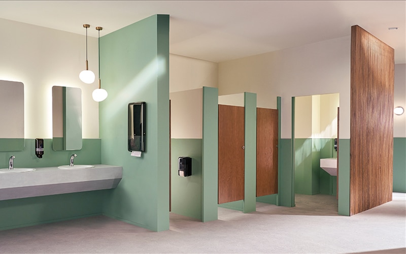 Des sanitaires modernes avec des murs vert clair et des portes de cabine de toilette en bois, avec deux lavabos et des miroirs, et des distributeurs de savon. Des distributeurs d’essuie-mains en papier et de désodorisant sont fixés au mur. Des sanitaires modernes avec des murs vert clair et des portes de cabine de toilette en bois, avec deux lavabos et des miroirs, et des distributeurs de savon. Des distributeurs d’essuie-mains en papier et de désodorisant sont fixés au mur.