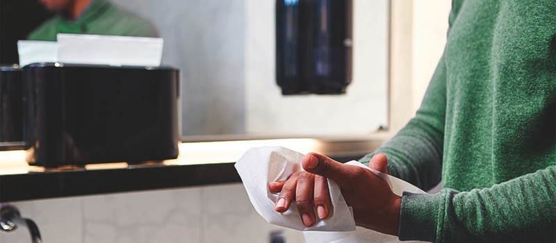 A man is wiping his hands with a paper towel, in the background he's in the mirror and there is also paper towel dispener and a soap dispenser.