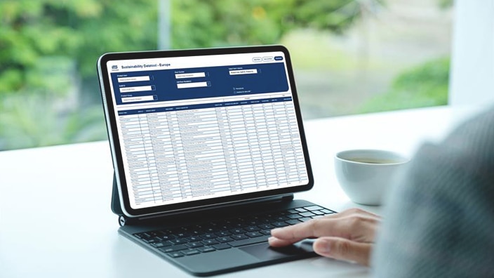 En person, der bruger en tablet med en datatabel åben på skærmen, med en kop på skrivebordet og grønt udenfor. En person, der bruger en tablet med en datatabel åben på skærmen, med en kop på skrivebordet og grønt udenfor.