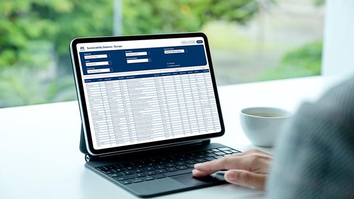 A person uses a tablet with a data table open on the screen, with a cup on the desk and greenery outside. A person uses a tablet with a data table open on the screen, with a cup on the desk and greenery outside.