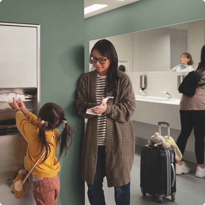Adult and child using a restroom area, with the child reaching for paper towels and luggage beside them Adult and child using a restroom area, with the child reaching for paper towels and luggage beside them