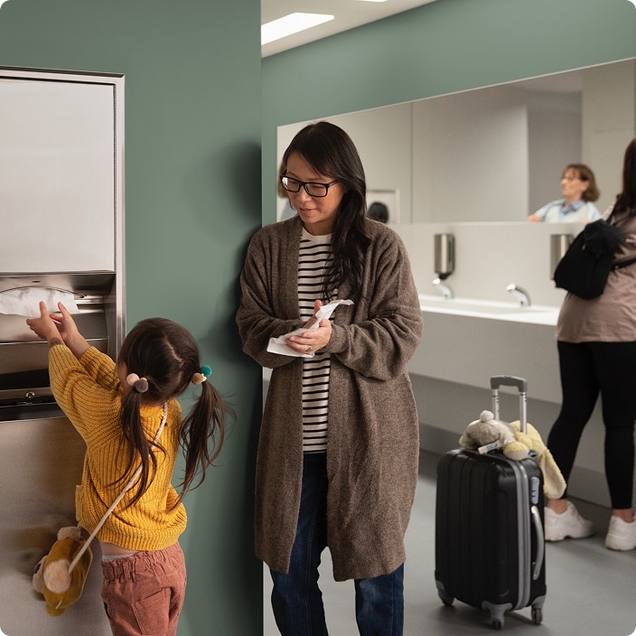 Adult and child using a restroom area, with the child reaching for paper towels and luggage beside them Adult and child using a restroom area, with the child reaching for paper towels and luggage beside them