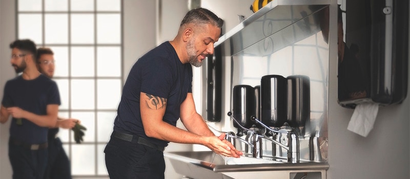 a man is washing his hands in a restroom with a long sink, several soap dispensers and paper towel dispensers, in the background to the left two more men