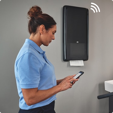 Cleaner checking refill information on a mobile phone next to a connected paper towel dispenser Cleaner checking refill information on a mobile phone next to a connected paper towel dispenser