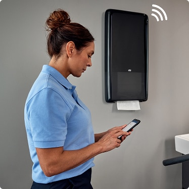 Cleaner checking refill information on a mobile phone next to a connected paper towel dispenser Cleaner checking refill information on a mobile phone next to a connected paper towel dispenser
