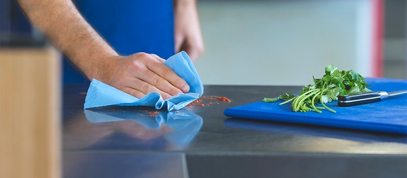 A hand is holding a blue cloth and is wiping some spills, in front a blue cutting board with fresh coriander and a knife
