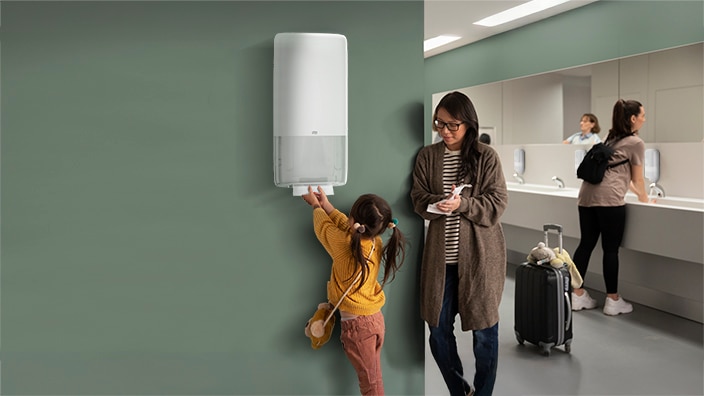 Kvinde står med et barn på et toilet og tager et papirhåndklæde fra en dispenser