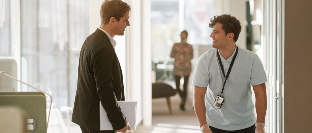 Two men stand in a bright hallway, smiling and talking while one holds a white box and the other wears a lanyard and gloves beside another box.