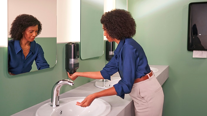A person using a wall‑mounted soap dispenser at a clean, modern bathroom sink with mirrors and hygiene equipment. A person using a wall‑mounted soap dispenser at a clean, modern bathroom sink with mirrors and hygiene equipment.