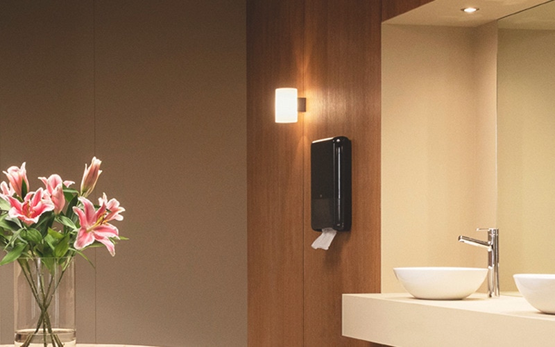 washroom with sink and black paper towel dispenser on the wall, to left a bouquet with flowers in a glass vase