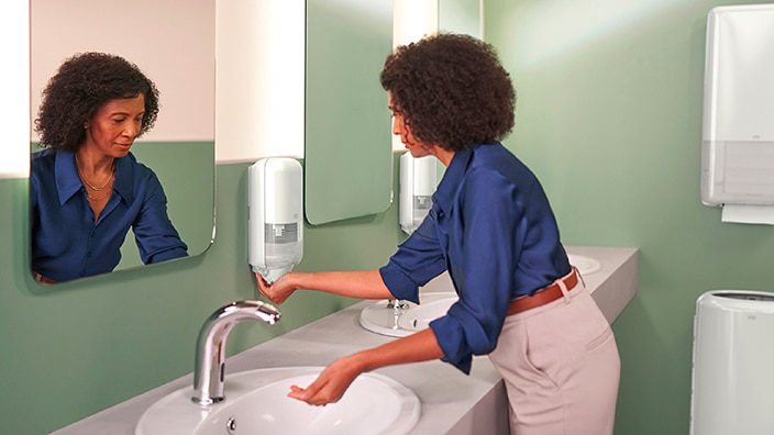 Eine Person benutzt einen wandmontierten Seifenspender an einem sauberen, modernen Waschbecken mit Spiegeln und Hygienesystemen.