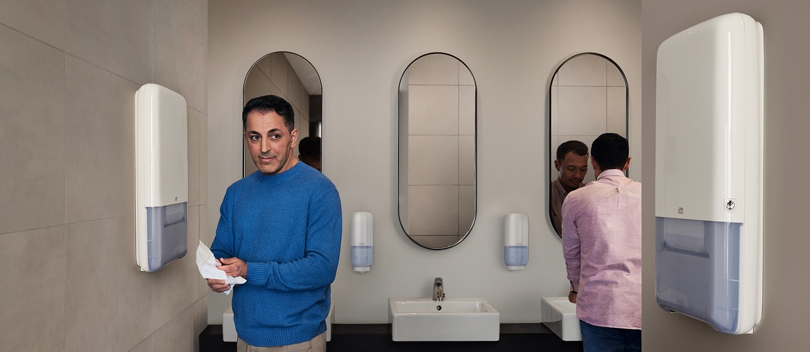 A person standing in a washroom drying their hands next to a white hand towel dispenser
