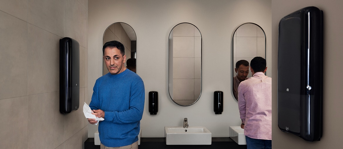 A person standing in a restroom drying their hands next to a black hand towel dispenser