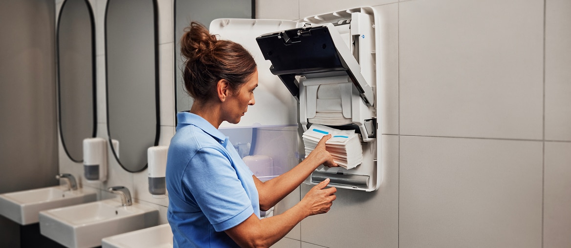A cleaner loading towels into a white electronic hand towel dispenser