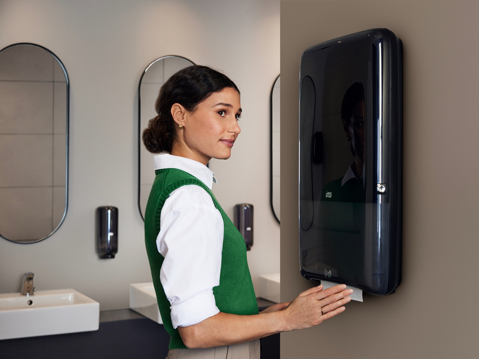 Woman using a black Tork PeakServe® Continuous™ paper hand towel dispenser mounted on a restroom wall. Woman using a black Tork PeakServe® Continuous™ paper hand towel dispenser mounted on a restroom wall.