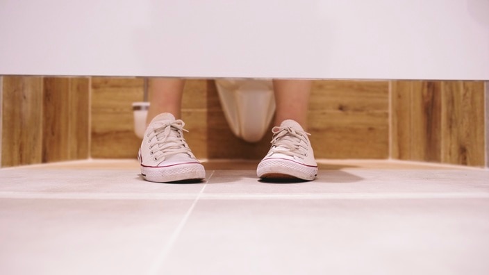 Beige sneakers with white soles seen from under a restroom stall door, with wooden paneling and tiled floor. Beige sneakers with white soles seen from under a restroom stall door, with wooden paneling and tiled floor.
