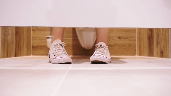 Beige sneakers with white soles seen from under a restroom stall door, with wooden paneling and tiled floor.